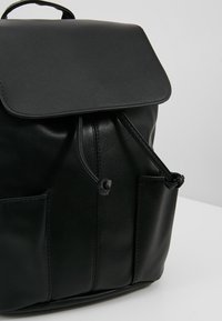 Černý kožený batoh s klopovým uzávěrem, stahovací šňůrkou nahoře, předními kapsami a hladkým povrchem. Obsahuje minimální dekorativní prvky.