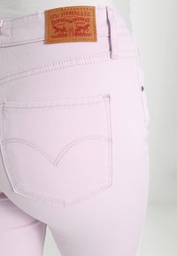 Jeans de mezclilla rosa claro con un bolsillo trasero que presenta un diseño de corazón bordado y una etiqueta de cuero marrón con detalles de marca en rojo.
