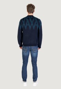 Homme de dos portant un pull marin avec un motif géométrique bleu, un jean bleu et des baskets blanches sur un fond clair.