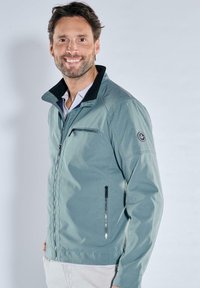 Hellgrüne Jacke mit Reißverschluss, schwarzem Kragen, Seitentaschen und Logo-Akzent am Ärmel. Glatte Textur und modernes Design.