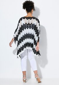 Svart och vit chevron-mönstrad poncho med fransdetaljer, tillverkad av ett öppet stickat tyg. Bärs över vita byxor och silverfärgade klackar.