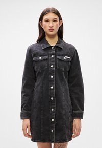 Vestido camiseiro em denim preto com frente abotoada, mangas longas, dois bolsos no peito e detalhes em botão prateado. A textura é resistente e suave.