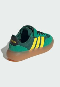 Grön och gul Adidas-sneaker med svarta snörningar, genomskinlig gumminubbe och logotyp på hälen, visad från baksidan mot en vit bakgrund.