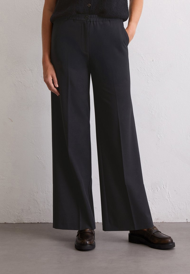 Pantalons larges noirs avec une texture lisse, des poches latérales et une fermeture à bouton à la taille. Portés avec des chaussures marron.