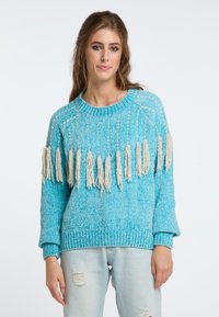 Maglione turchese lavorato a maglia con un motivo testurizzato e frange color crema lungo l'orlo. Abbinato a jeans chiari strappati di colore blu.
