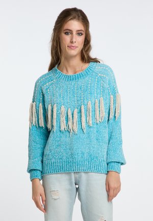 Türkiser Strickpullover mit einem strukturierten Muster und kremfarbigen Fransen am Saum. Kombiniert mit hellblauen, ausgebleichten Jeans.