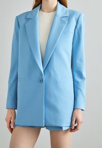 Blazer azzurro chiaro dal design sartoriale, presenta un rever a punte, chiusura con un solo bottone e due tasche frontali. Texture liscia.