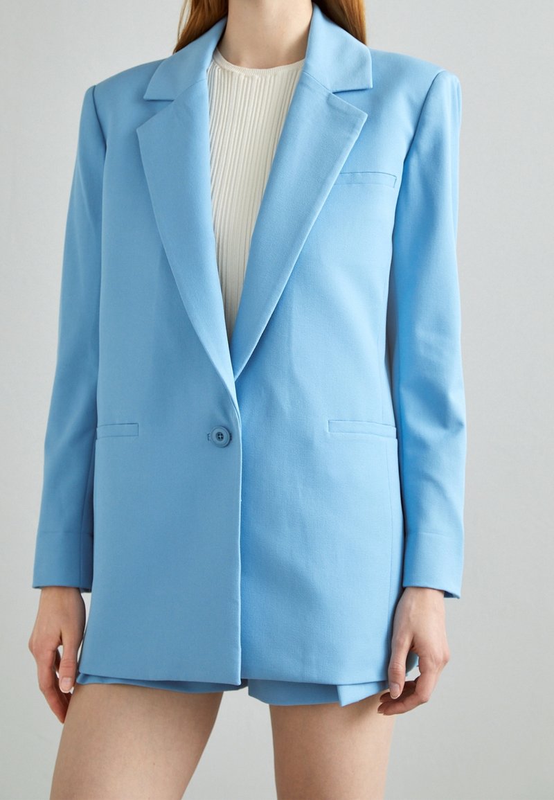 Blazer azzurro chiaro dal design sartoriale, presenta un rever a punte, chiusura con un solo bottone e due tasche frontali. Texture liscia.