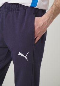 Námornícke modré športové nohavice vyrobené z mäkkého materiálu, s elastickým pásom a bielym logom Puma na ľavom stehne. Viditeľná bočná vrecko.