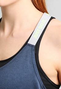 Sportbh med svart och mörkblåt tyg, med en platt halsringning och breda remmar. Remmarna har texten "JUST DO IT" i reflekterande ljusgrön färg.