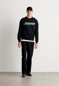 Man walking wearing black cap, black sweatshirt with blue-green text, black pants, and black shoes on plain gray floor and white wall background.
