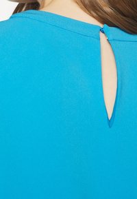 Modrá blůza s kulatým výstřihem a klíčovou dírkou na zadní straně. Hladký povrch a lehká tkanina.