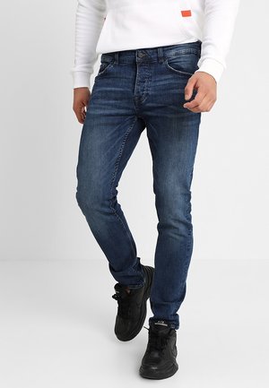 Homme portant un jean bleu foncé slim, des chaussures de sport noires et un haut blanc à manches longues, debout devant un fond blanc uni.