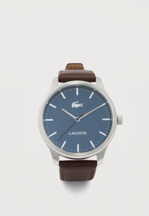 Montre ronde en argent avec cadran bleu, index blancs, aiguilles argentées, logo Lacoste, et bracelet en cuir marron sur fond blanc.