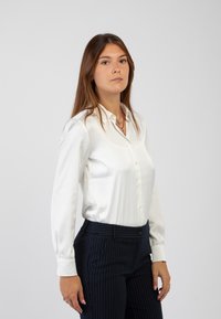 Camicia button-up bianca in satin con maniche lunghe, abbinata a pantaloni scuri a righe sottili. Texture liscia e vestibilità su misura.