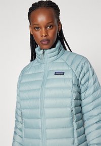 Svijetloplava puffer jakna s vertikalnim kvadratnim dizajnom, visokim ovratnikom i džepovima na zip. Sadrži Patagonia logo s lijeve strane.