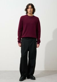 Burgundin villane sviiter, millel on ümmargune kaelus, koos laiade mustade pükste ja mustade lipsudega kingadega, seistes ühetaolise tausta ees.