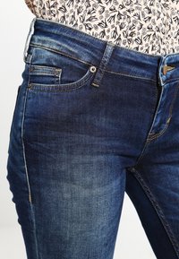 Jeans azul oscuro hechos de denim texturizado. Las características incluyen un diseño clásico de cinco bolsillos, un sutil desvanecimiento y acentos de hardware en tono cobre.
