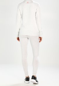 Persona con un top a maniche lunghe grigio chiaro e leggings con logo Adidas rosa, in piedi di spalle, con sneaker bianche e nere.