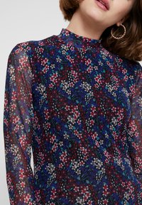 Vrouw draagt een transparante top met hoge hals en lange mouwen, bedrukt met een dicht bloemmotief in rood, blauw, wit en groen, en grote, gestructureerde oorringen.