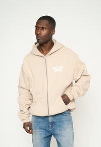 Sudadera beige con cremallera hecha de una tela suave, que presenta una gran capucha, bolsillos frontales y texto impreso en el lado izquierdo del pecho. Combinada con jeans azules.