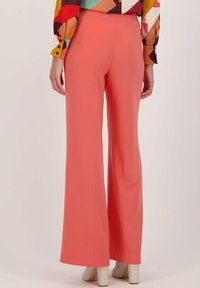 Koraalkleurige wijd uiteindigende broek van een gladde stof, met een tailleband en een vloeiende silhoeet. Gepaard met beige hoge hakken.