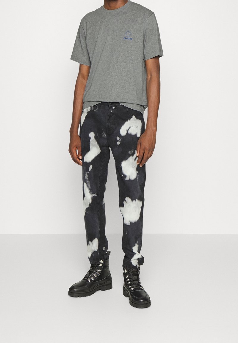 T-shirt gris à manches courtes avec logo bleu, associé à un jean noir orné de motifs blancs ressemblant à des nuages ; des bottes noires à lacets complètent la tenue.