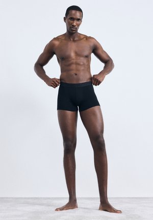 Čierne boxerky vyrobené z mäkkej tkaniny s vzorovaným elastickým pásom. Model stojí naboso na svetlosivom pozadí.