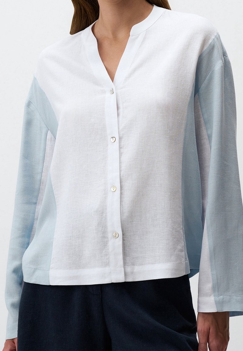 Femme portant une chemise à manches longues avec devant blanc et manches et côtés bleu clair, assortie à un pantalon foncé.