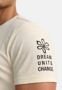 Cremefarbenes Kurzarm-T-Shirt mit strukturierter Oberfläche, das schwarzen Drucktext und eine Blumen-Grafik auf dem linken Ärmel zeigt.