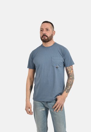 Uomo con barba e tatuaggi che indossa una maglietta blu con tasche e jeans azzurri, in piedi davanti a uno sfondo chiaro uniforme.