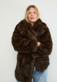Cappotto in finta pelliccia marrone con una texture soffice, colletto oversize e cuciture diagonali. Indossato sopra un outfit in denim azzurro chiaro.