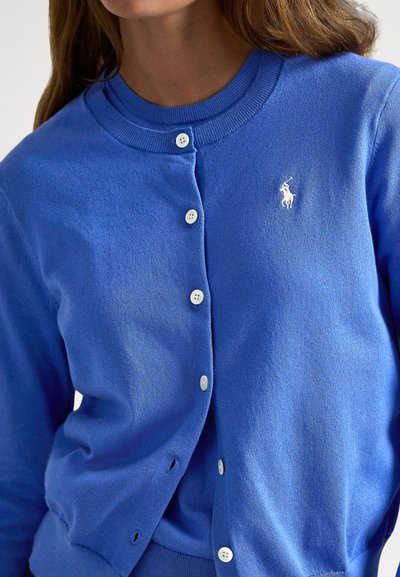 Μπλε πλεκτή ζακέτα με στρογγυλή λαιμόκοψη, πέντε λευκά κουμπιά και μικρό κ�εντημένο λογότυπο στο αριστερό στήθος. Μαλακή υφή.