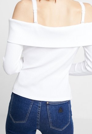Femme portant un haut blanc côtelé à épaules dénudées avec une bretelle de soutien-gorge blanche visible et un jean en denim bleu foncé vu de dos.