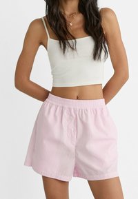 Femme portant un top court blanc et un short élastique rayé rose clair, se tenant avec les mains derrière le dos.