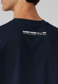 Granatowy sweter crewneck z teksturowanym wykończeniem. Białe drukowane napisy z tyłu brzmią "HANGING AROUND Since 1950" z informacjami o prawach autorskich.