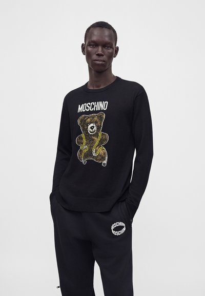 Jeune homme portant un pull noir Moschino avec un motif d'ourson et un pantalon noir avec un logo Moschino blanc sur la poche gauche.