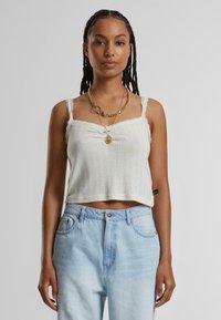 Vit crop top med strukturerad stickning, spetsdetaljer på axelbanden och en märkesetikett i nederkant, kombinerad med ljusblå högmidjade jeans.