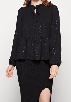 Kobieta ubrana w czarną bluzkę z długim rękawem z srebrnymi kropkami i wiązaniem przy dekolcie, zestawioną z czarną prążkowaną dzianinową spódnicą z przednim rozcięciem.