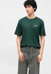 Camiseta de manga corta de color verde oscuro en tejido suave, con un pequeño logo blanco en el pecho. Corte holgado combinada con pantalones gris claro.