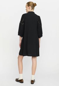 Schwarzes knielanges Kleid mit Puffärmeln, Spitzenakzenten an den Schultern und Manschetten sowie einer lockeren Passform. Kombiniert mit braunen Schuhen und weißen Socken.