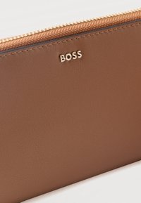 Portefeuille en cuir marron avec fermeture éclair dorée et petit logo "BOSS" doré sur le devant.