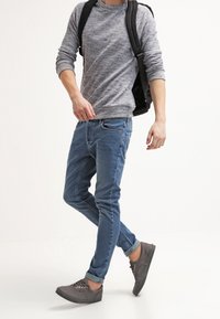 Suéter gris, pantalones vaqueros azules y zapatillas deportivas grises. El conjunto presenta un estilo casual con un suéter texturizado y vaqueros de corte ajustado. Mochila negra llevada.