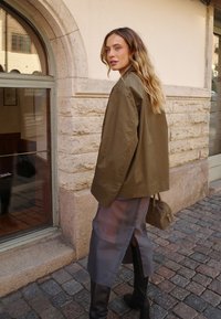 Olivenfarvet oversized jakke, gennemsigtig grå nederdel med sideslids, sorte knæhøje støvler og lille beige håndtaske, sat mod brostensbelagt gade.