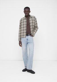 Koszula z flaneli w zielono-beżową kratę na brązowym t-shircie, zestawiona z jasnoniebieskimi prostymi dżinsami i czarnymi tenisówkami.