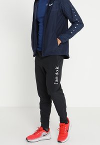 Person in schwarzen Nike Jogginghosen mit "Just do it."-Aufdruck, navyblauer Nike-Jacke und leuchtend roten Nike-Laufschuhen. Hände in den Taschen.