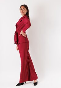 Completo a due pezzi in un profondo bordeaux, composto da un blazer sartoriale con cintura in vita e pantaloni a gamba larga. Tessuto liscio abbinato a tacchi a spillo neri.