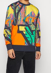 Sweatshirt colorée présentant un motif géométrique vibrant en jaune, orange, vert et bleu avec des accents noirs. Poignets et ourlet côtelés.