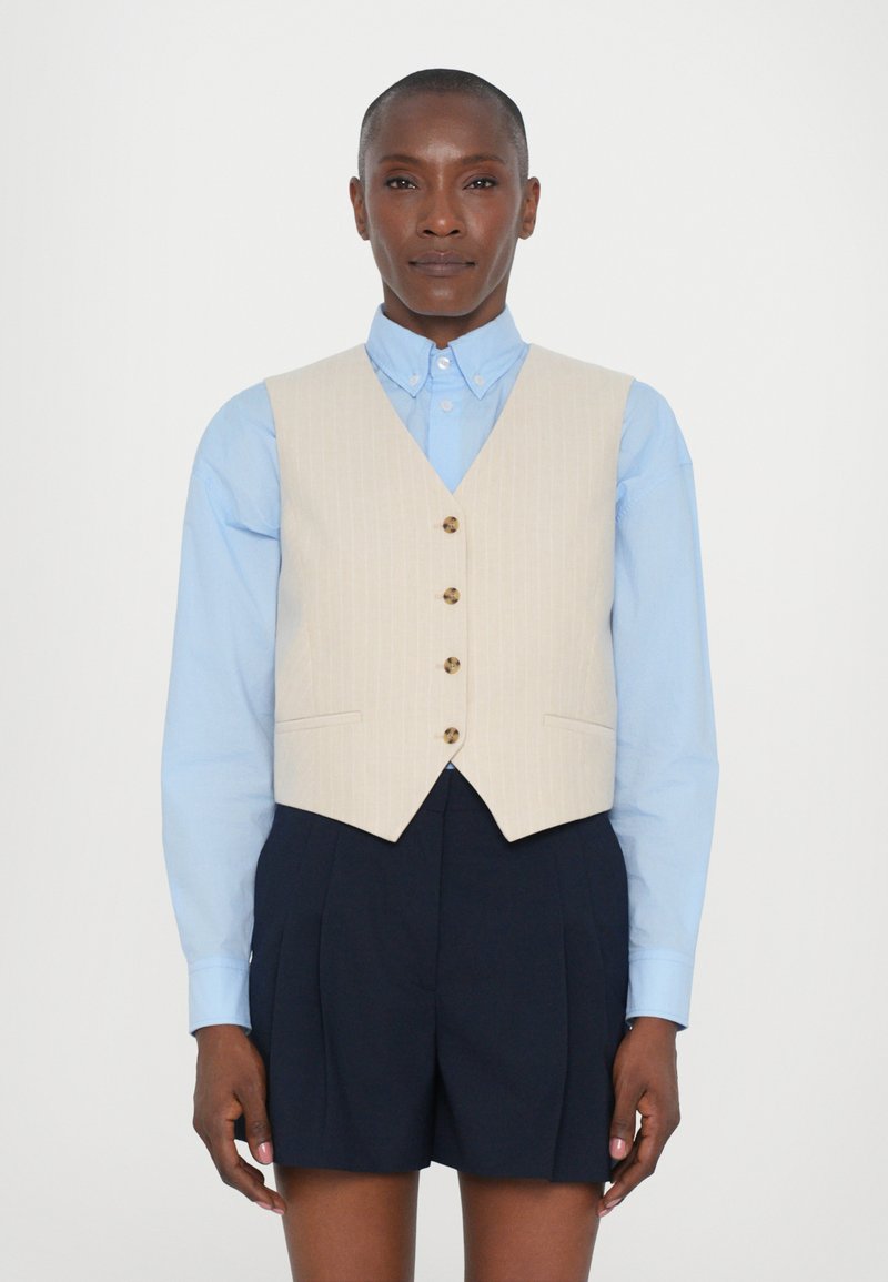 Beige pinstripe liivi, jossa on viisi ruskeaa nappia, käytetään vaaleansinisellä kauluspaidalla ja paritetaan tummansiniharmaiden shortsien kanssa. Sileä kangastekstuuri.