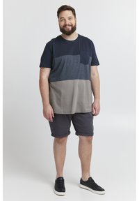 Marineblå og grå fargeblokkerte T-skjorter med lomme, paret med mørkegrå shorts. Modellen har på seg svarte joggesko. Enkel bakgrunn.
